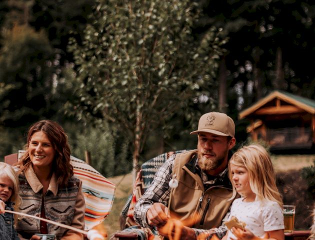 A family is gathered around a campfire in a forested area, roasting marshmallows. There's a cozy cabin in the background.