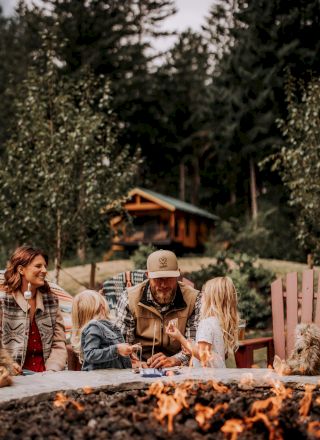 A family sitting around a fire pit outdoors, with wooded hills and a cabin in the background.