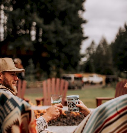 Two people wrapped in patterned blankets clinking mugs outdoors, with trees in the background.