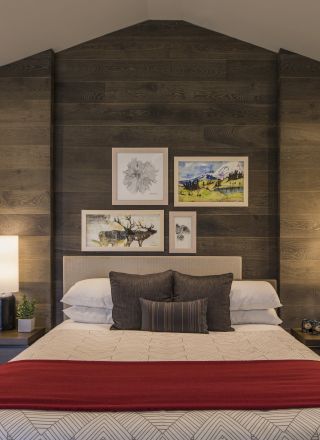 A cozy bedroom with a bed, red blanket, wooden accent wall, framed art, lamps, and a desk with a microwave and deer head decor.