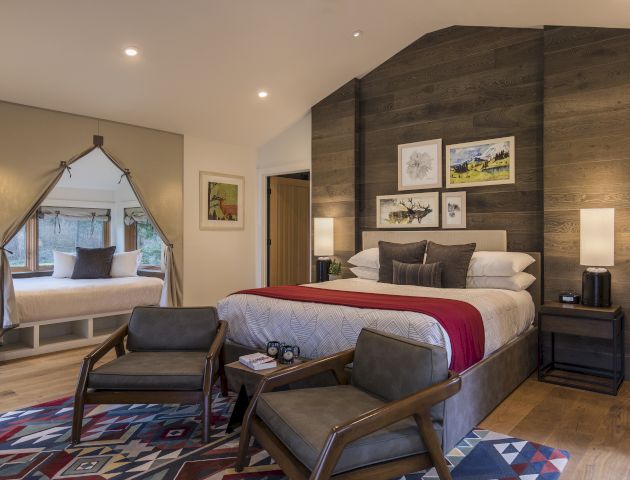 A cozy bedroom with a double bed, two chairs, a rug, and a nook with a window seat, decorated in warm tones and wooden accents.