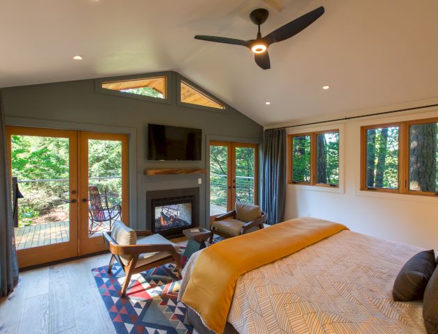 A cozy bedroom features a bed, fireplace, TV, and chairs, with large windows offering a view of the forest outside, and a ceiling fan.