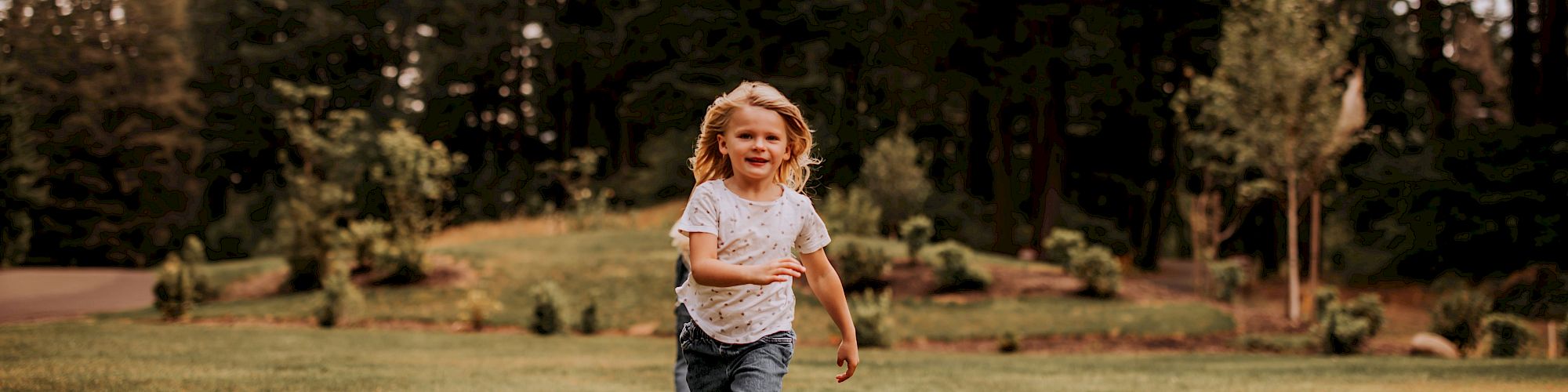 A child runs joyfully on a grassy field surrounded by tall trees and a cloudy sky, exuding a sense of freedom and happiness.