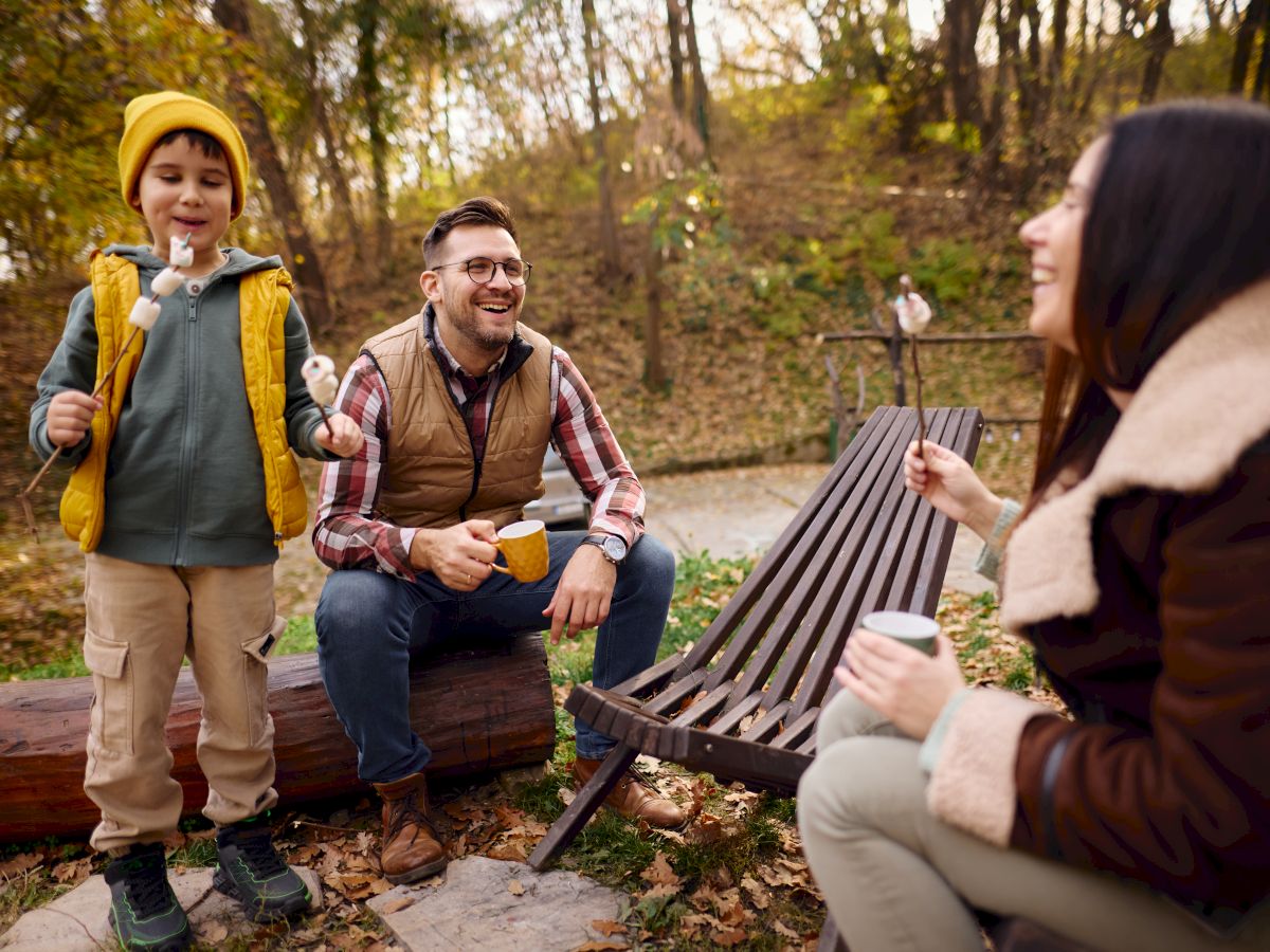 A family is sitting outdoors, enjoying roasted marshmallows and hot drinks. They seem to be having a pleasant time in a wooded area.