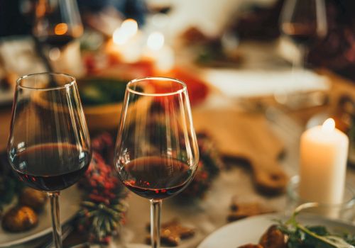 Two glasses of red wine are on a table with food, candles, and festive decorations, creating a warm, cozy atmosphere.