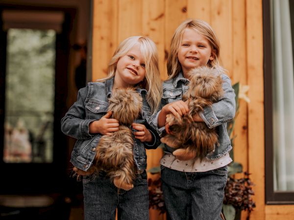 Two children in denim outfits stand in front of a wooden house, each holding a small, fluffy dog, smiling for the camera.
