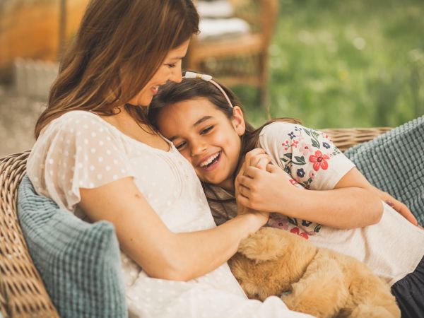 Two women share a tender moment on a wicker sofa, one cuddling a child and a fluffy dog, a cozy, loving family scene.