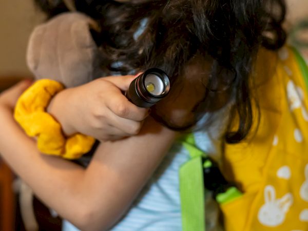 A child hugs a white teddy bear while shining a small flashlight on their shoulder, wearing a yellow backpack with bunny designs.