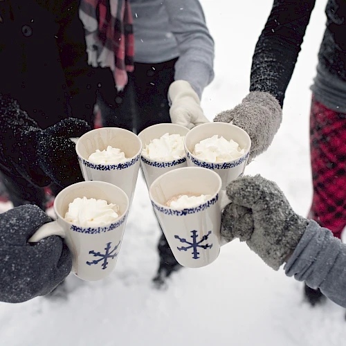 Five people standing in snow clink mugs filled with whipped cream, wearing cozy winter clothing and gloves.
