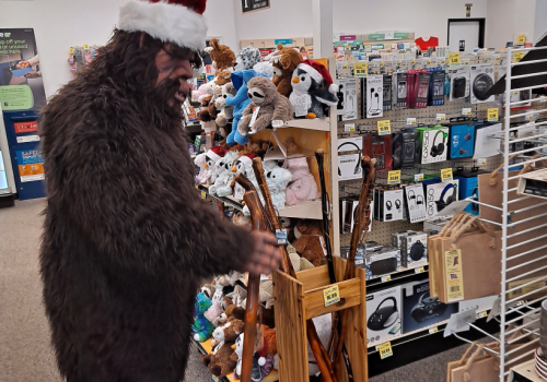 A person in a Bigfoot costume with a Santa hat shops in a store aisle filled with various items, including toys and walking sticks.