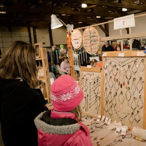 People are browsing jewelry at a market stall. The display includes necklaces and bracelets on a wooden stand, indoors.