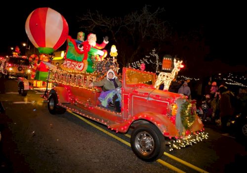 A festive, brightly lit parade float with Christmas decorations, including a reindeer, Santa, and a large balloon on a classic red vehicle.