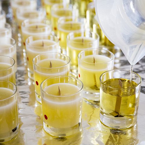 Rows of glass containers with yellow wax and wicks, with liquid being poured into one of them, likely for candle making.
