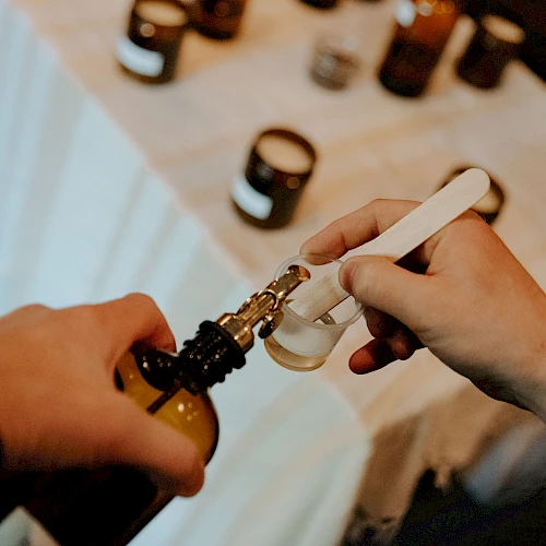 A person is filling a small container with liquid using a dropper, surrounded by several small jars on a table.