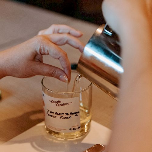 A person is pouring liquid from a metal pitcher into a glass with text on it, possibly preparing a drink on a table.
