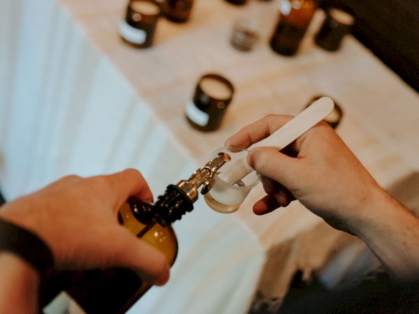 A person is pouring liquid into a small bottle using a dropper. Candles are visible on a table in the background.