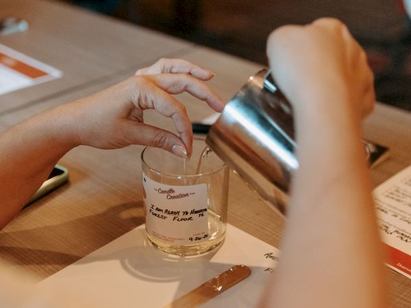 A person is pouring liquid from a metal container into a glass with a wick, possibly making a candle, over a table with papers.