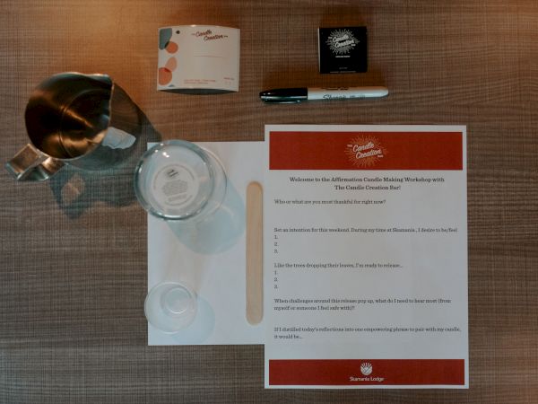 The image shows a table setting with a metallic jug, glass with coaster, notepad, pen, matchbox, and a piece of paper with some text.