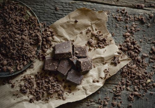 The image shows pieces of chocolate on parchment paper, surrounded by chocolate shavings on a rustic wooden surface.