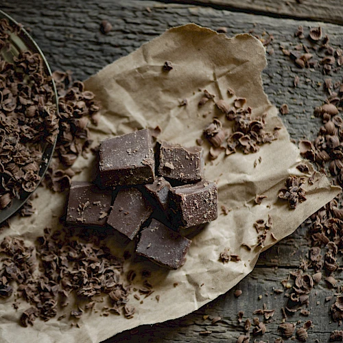 The image shows pieces of chocolate on parchment paper, surrounded by chocolate shavings on a rustic wooden surface.