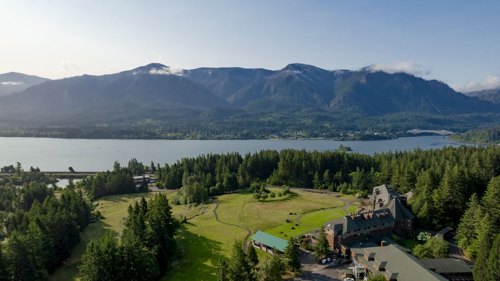 A scenic landscape with a lake, forest, and building. Mountains rise in the background, under a clear sky.