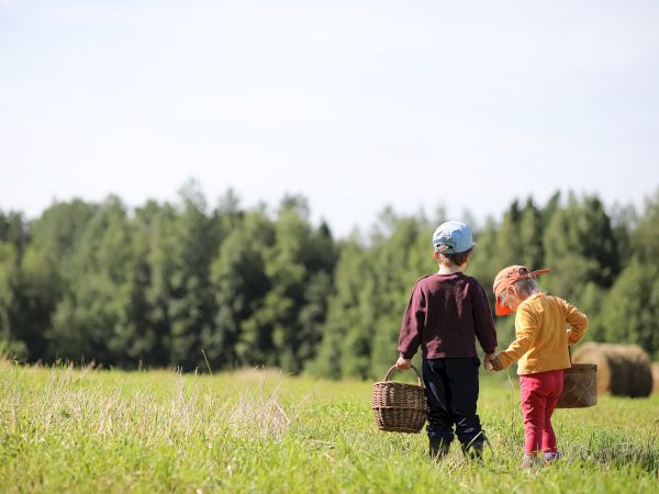 Two children in a field carry baskets, surrounded by greenery and hay bales under a clear sky.
