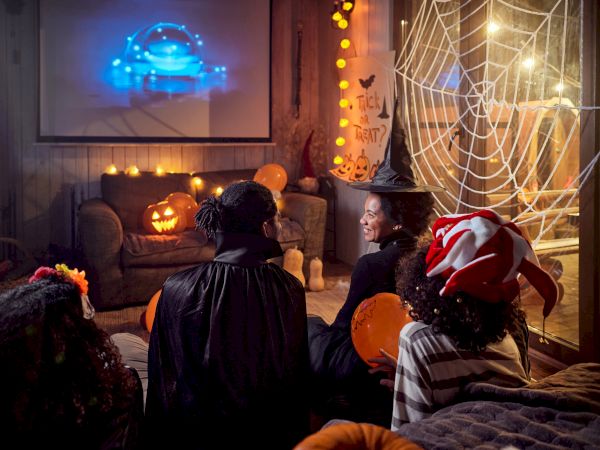 People in costumes watching a movie in a Halloween-themed room with pumpkin decorations and a cobweb.