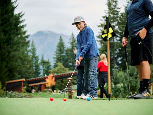 A child and an adult play mini-golf on a course surrounded by trees and mountains, with colorful golf balls and a flag in view.